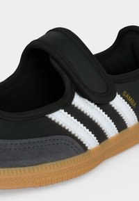 Μαύρο παπούτσι Adidas Samba με λευκές ρίγες, καουτσούκ σόλα και φαρδιά κλείσιμο με Velcro.