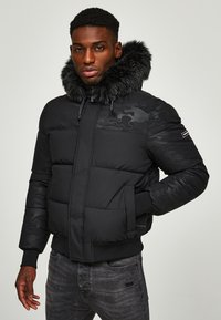 Chaqueta de plumas negra con capucha forrada de pelo, que presenta un patrón de camuflaje en los hombros, cremallera frontal y puños y dobladillo acanalados.
