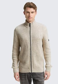 Beige gebreide trui met een hoge kraag, voorzien van een zwarte rits en ribbelstructuur. Opvallende spikkels voegen visuele interesse toe.