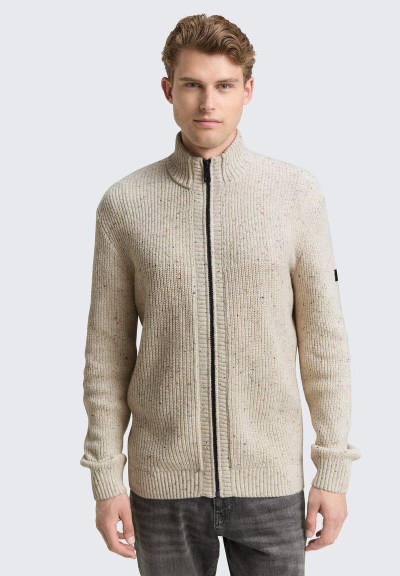 Beige gebreide trui met een hoge kraag, voorzien van een zwarte rits en ribbelstructuur. Opvallende spikkels voegen visuele interesse toe.