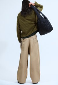 Personne avec de longs cheveux noirs portant un pull olive et un pantalon beige large, tenant un grand sac noir matelassé sur l'épaule.