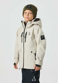 Beige waterproof jacket with a hood, black zipper, and multiple front pockets. Features logo patches and black accents. Paired with black pants and a green beanie.