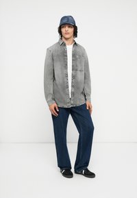 Grijze denim shirt met knoopsluiting aan de voorkant en een borstzak, gecombineerd met donkerblauwe wijde spijkerbroek en een blauwe met patroon bedekte pet. Zwarte sneakers maken de look compleet.