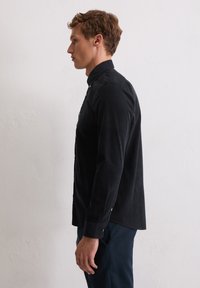 Schwarzes Knopfhemd mit hohem Kragen, langen Ärmeln und Stoffstruktur, versehen mit Metallknopfakzenten und einem geschwungenen Saum.