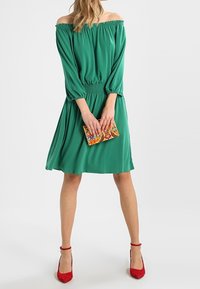 Zielona sukienka bez ramiączek z elastyczną talią, swobodnym krojem i trzema ćwierć rękawami. Połączona z kolorową torebką w wzory i czerwonymi szpilkami.