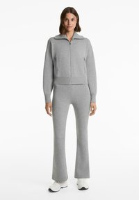 Vrouw in een bijpassende grijze zip-up sweater en uitlopende broek met witte sneakers, staand tegen een effen witte achtergrond.