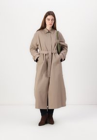 Bežični trench plašč z pasom, dolgimi rokavi in ovratnikom. Model pozira s rokami v žepih, oblečen v temne kavbojke in rjave škornje.