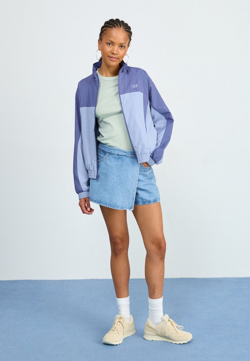 Cazadora ligera, a rayas moradas y azules, con cierre de cremallera, combinada con una camiseta verde claro y una falda de mezclilla. Zapatillas completan el look.