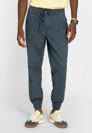 Hombre con pantalones jogger de puño elástico azul marino, zapatillas amarillas y blancas, camiseta amarilla, reloj de pulsera y pulsera, posando contra un fondo blanco.