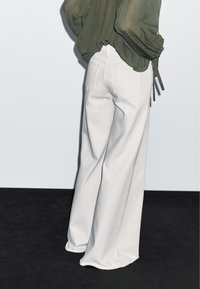 Jeans bianchi a gamba larga con vita alta, orlo rifinito e due tasche posteriori, abbinati a una blusa leggera, verde oliva, arricciata.