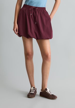 Femme portant une jupe boule bordeaux à taille élastique avec un cordon de serrage et des baskets marron, debout devant un fond gris uni.