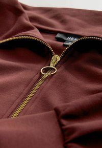 Tamsiai raudonas užtraukiamas džemperis su auksinės spalvos užtrauktuku ir apvaliu užtrauktuko užtrauktuku. Minkšta medžiaga, pasižymi siūlėmis ir viduje apykaklėje matoma etiketė.