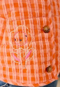 Orange checkered fabric with stitched smiling flower design and two visible brown buttons on a quilted garment.