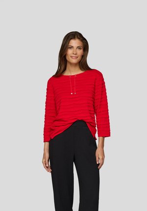 Frau in einem strukturierten roten Langarmoberteil mit Kordelzügen und schwarzen hochtaillierten Hosen, die vor einem schlichten grauen Hintergrund steht.