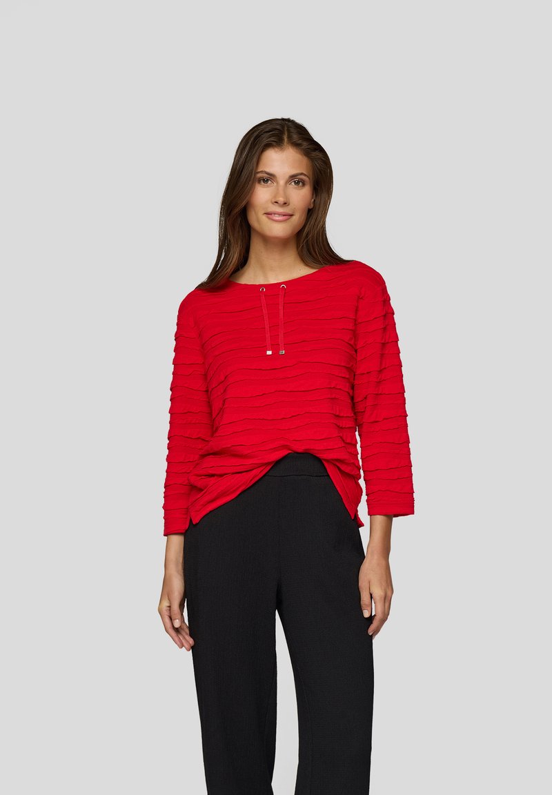 Frau in einem strukturierten roten Langarmoberteil mit Kordelzügen und schwarzen hochtaillierten Hosen, die vor einem schlichten grauen Hintergrund steht.