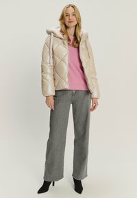 Cappotto morbido di un beige chiaro con un motivo trapuntato a rombi e cappuccio. Indossato sopra un maglione rosa e abbinato a jeans grigi e tacchi neri.