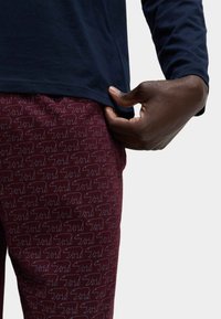 Pantalones color burdeos con un estampado de osos en todo el tejido en un color claro en contraste, combinados con una camiseta de manga larga azul marino. Tejido suave y diseño ajustado.