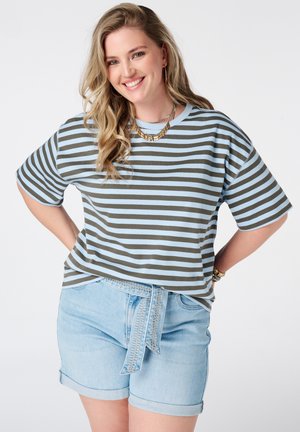 Lachende vrouw met lang blond haar, die een blauw-grijs gestreept shirt, lichte denimshorts met riem en gouden sieraden draagt.