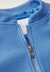 Blå fleecjacka med hög krage och silver dragkedja. Slät textur med enfärgad design; inre foder är en ljusare blå.
