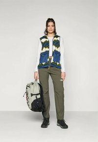 Zils un zaļš rakstains flīsa veste ar divām priekšējām kabatām, apvienota ar olīvu zaļām biksēm un melniem pārgājienu apaviem, turēt pelēku mugursomu.