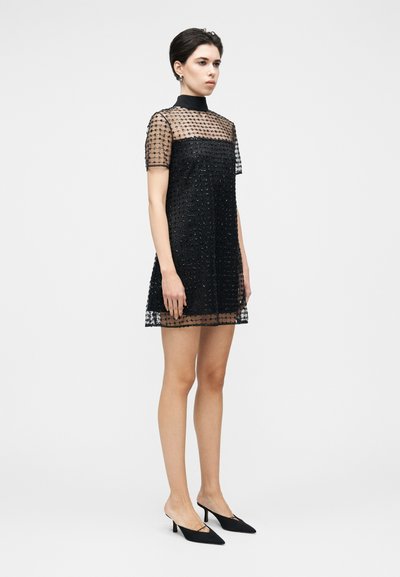 Black mesh dress with short sleeves, featuring intricate bead detailing and a high neck. Paired with sleek black heeled mules.