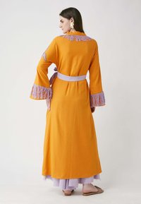 Largo vestido naranja con flecos lavanda en las mangas y el cuello, ceñido con un cinturón de color púrpura claro; falda de lila en capas visible por debajo.