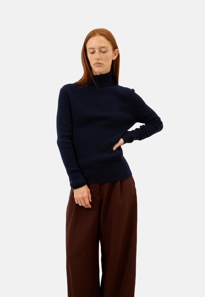 Maglione a collo alto blu navy realizzato in morbido tessuto a maglia, silhouette aderente e maniche lunghe. Abbinato a pantaloni larghi marroni.