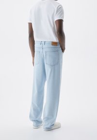 Jeans de mezclilla azul claro con un corte relajado, que cuentan con dos bolsillos traseros y un parche de cuero con el logo cerca de la cintura. Combinados con una camiseta blanca.