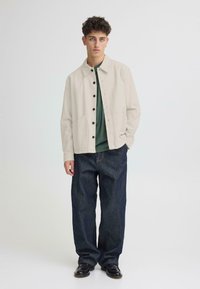 Veste en coton beige clair avec des boutons noirs, deux poches avant, portée sur une chemise vert foncé, associée à un pantalon en denim foncé ample.