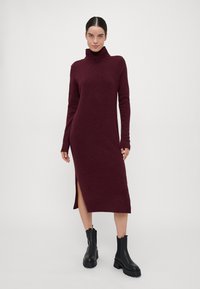 Burgundy kötött ruha magas nyakú, hosszú ujjú, oldalsó hasítékkal és bordás mandzsettákkal; fekete bokacsizmával viselve, semleges háttér előtt.