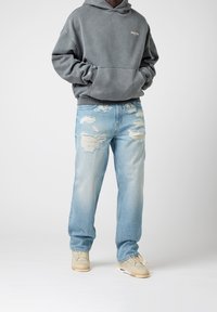 Sudadera gris con un bolsillo en la parte frontal, combinada con pantalones vaqueros desgastados de lavado claro que muestran grandes desgarros. Zapatillas beige completan el conjunto.