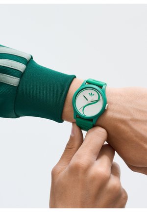 Zelena silikonska ura na zapestju s belim številčnico, ki se ponaša z minimalističnim dizajnom in vzorcem, podoben listu; nosi se na roki, ki prilagaja zapestnico.