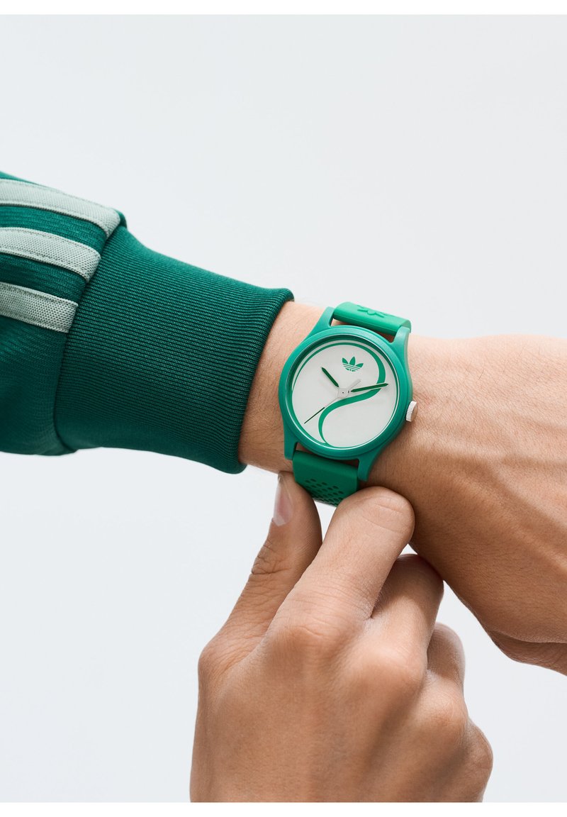 Roheline silikoonist randmekell valge kellapinnaga, minimalistliku disainiga ja lehekujundusega; kantud käel, mis reguleerib rihma.