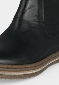 Gros plan sur une botte Chelsea en cuir noir avec un panneau latéral élastique et une semelle en caoutchouc marron superposée.