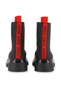 Botins pretos até ao tornozelo feitos de material sintético liso com sola preta grossa. Apresentam riscas vermelhas com o logótipo na parte de trás para visibilidade e identificação da marca.