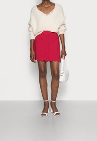 Neuleva cream-coloured paita V-kaula-aukolla, yhdistettynä punaiseen hameeseen, jossa on aaltoileva helma ja nappikiinnitys. Valkoiset hihnatossut ja käsilaukku.