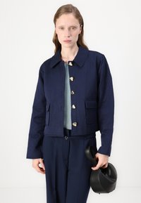 Marineblauwe cropped jack met een kraag, hartvormige gouden knopen, twee voorzakken en een gestructureerde stof. Gecombineerd met een groene trui en een zwarte tas.