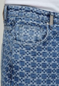 Jeans de denim con un patrón de tablero de ajedrez azul, diseño de cinco bolsillos y acentos de herrajes metálicos. Superficie texturizada con tonos contrastantes.