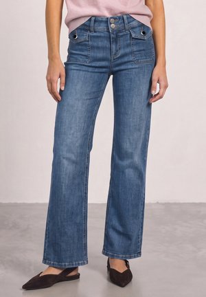 Femme portant un jean bleu taille haute à jambes larges avec poches rabat à l'avant et fermeture à double bouton, associé à des ballerines noires pointues.