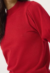 Czerwona, dziana sweter o wysokim okrągłym dekolcie i bufiastych rękawach, o lekko błyszczącej teksturze i dopasowanym kroju.