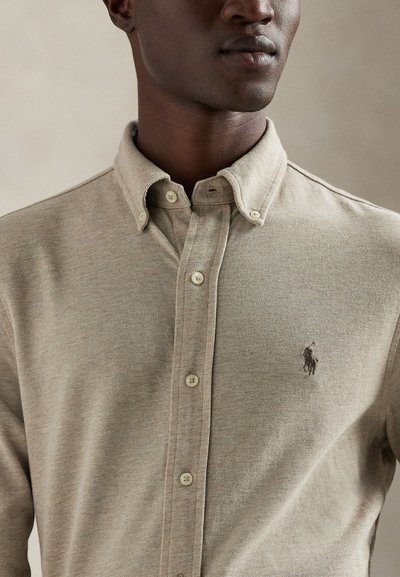 Camisa bege com gola, feita de tecido texturizado com fecho de botões, apresentando um pequeno logótipo bordado no lado esquerdo do peito.