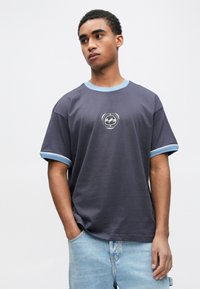 Νέος άνδρας που φοράει μπλούζα σε σκούρο μπλε με ανοιχτό μπλε περίγραμμα και μικρό λευκό αφηρημένο λογότυπο, συνδυασμένη με ανοιχτό μπλε τζιν σορτς, στέκεται με το χέρι στην τσέπη.