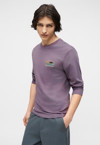 Νέος άνδρας με σκούρα μαλλιά που φοράει μωβ πουκάμισο μακρυμάνικο με μικρό πολύχρωμο λογότυπο Billabong στο στήθος, με τα χέρια στις τσέπες του μπλε-γκρι παντελονιού.