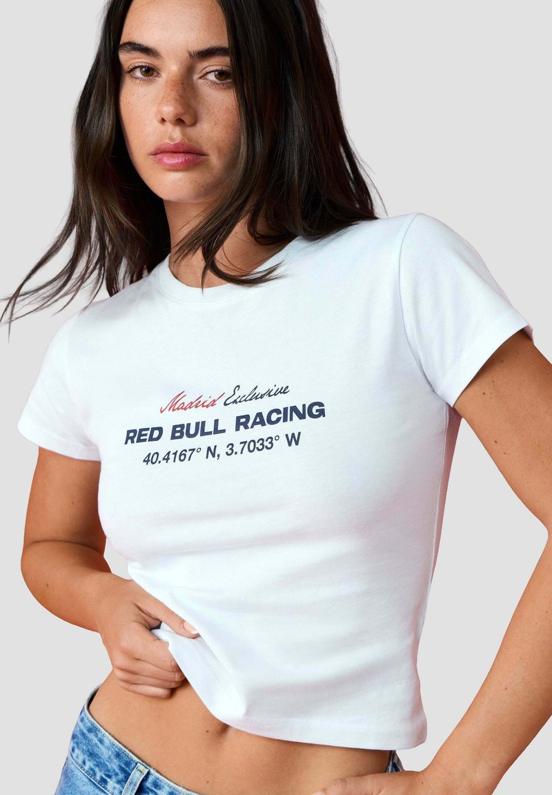Young woman with dark hair wearing a white cropped T-shirt with "Madrid Exclusive RED BULL RACING" and coordinates printed on front.