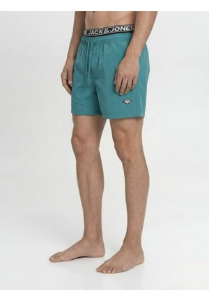 Mężczyzna w turkusowych szortach kąpielowych stoi boso, pokazując elastyczny pas z czarno-białym logo Jack & Jones.