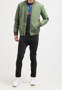 Uomo che indossa una giacca bomber verde oliva, una camicia floreale blu navy, jeans strappati neri e sneakers bianche, in piedi con una mano in tasca.