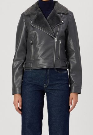 Person iført en sort jakke af kunstlæder med bred shearlingkrave, asymmetrisk lynlås og mørkeblå jeans mod en ensfarvet baggrund.