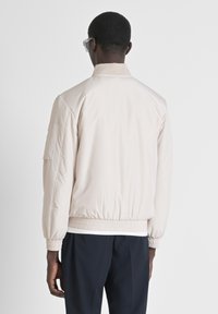Bomber jacket de un beige claro con un cuello y puños de canalé, cinturilla elástica y un bolsillo lateral. Textura de tela suave y diseño sencillo.