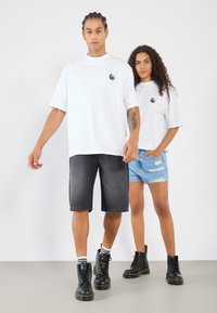 To personer står foran en hvid baggrund. Den mandlige bærer en hvid oversized t-shirt og mørke denim shorts; den kvindelige har en hvid t-shirt og slidte blå shorts på. Begge har sorte combat støvler på.