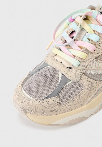 Μπεζ αθλητικό παπούτσι με περίτεχνα σχέδια, πάνελ από δίχτυ, κορδόνια σε παστέλ χρώματα με χάντρες σε ανοιχτό γκρι φόντο.
